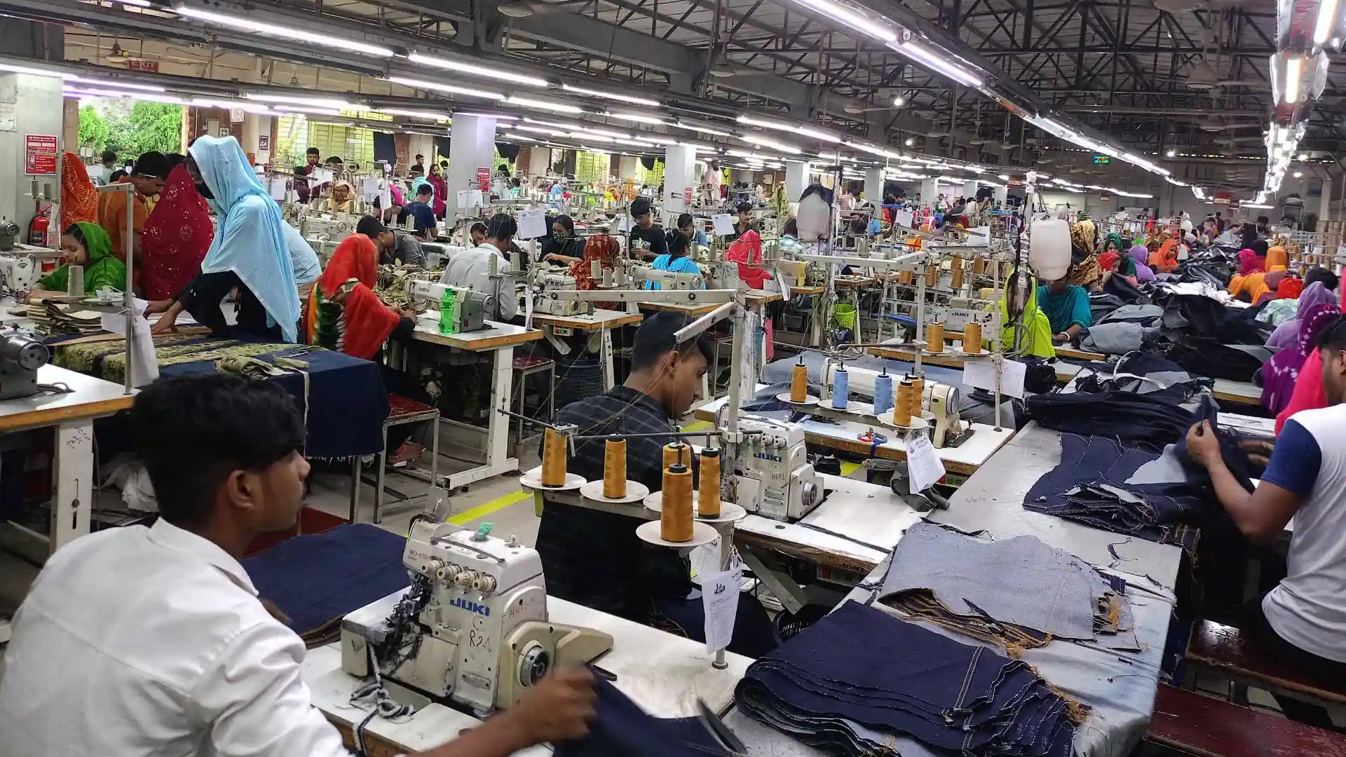 Workers sewing denim garments on the production floor with rows of industrial sewing machines in operation.