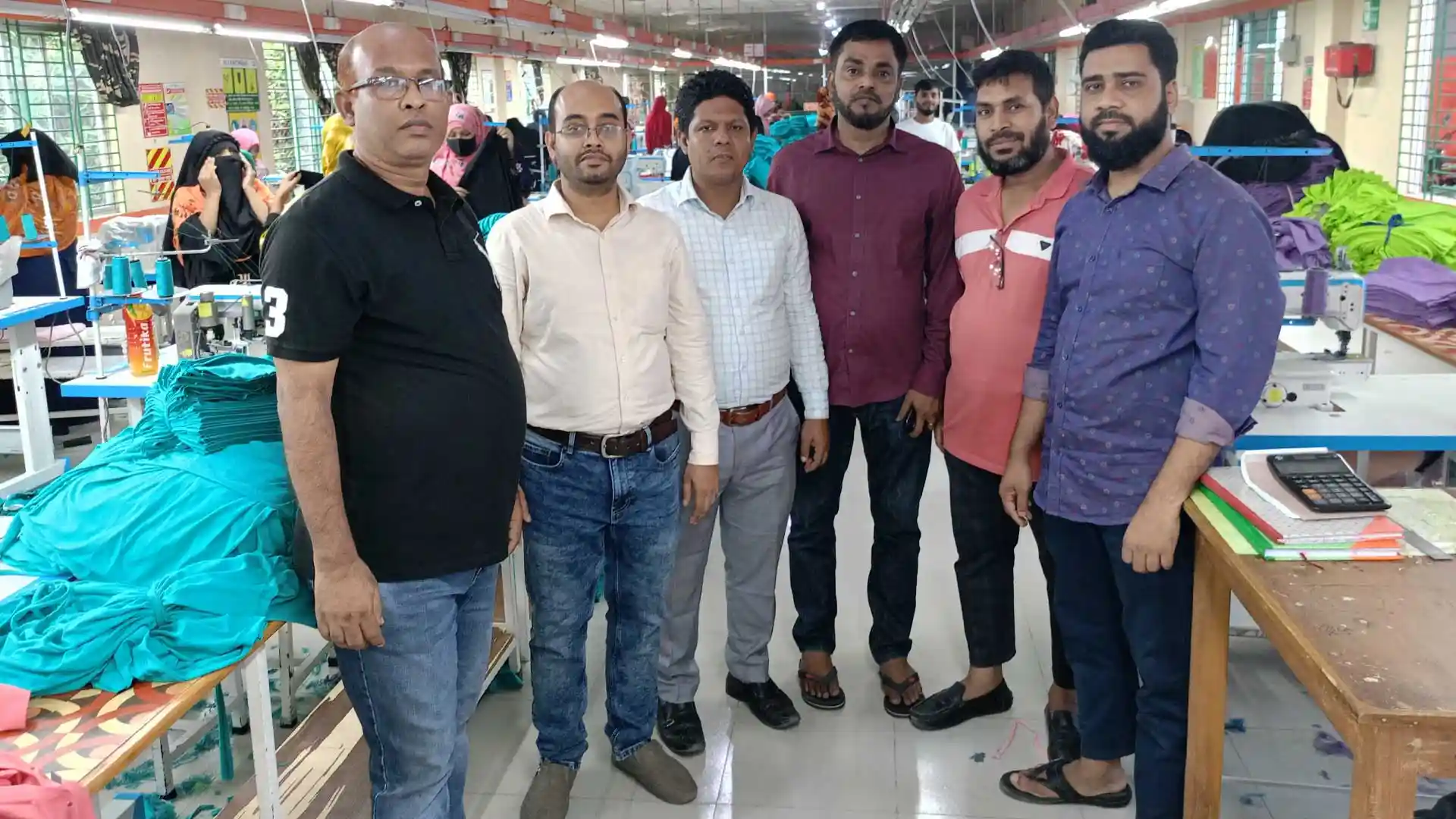 RYZEAL Sourcing team visiting a garment factory floor, standing together while workers are engaged in apparel production in the background.