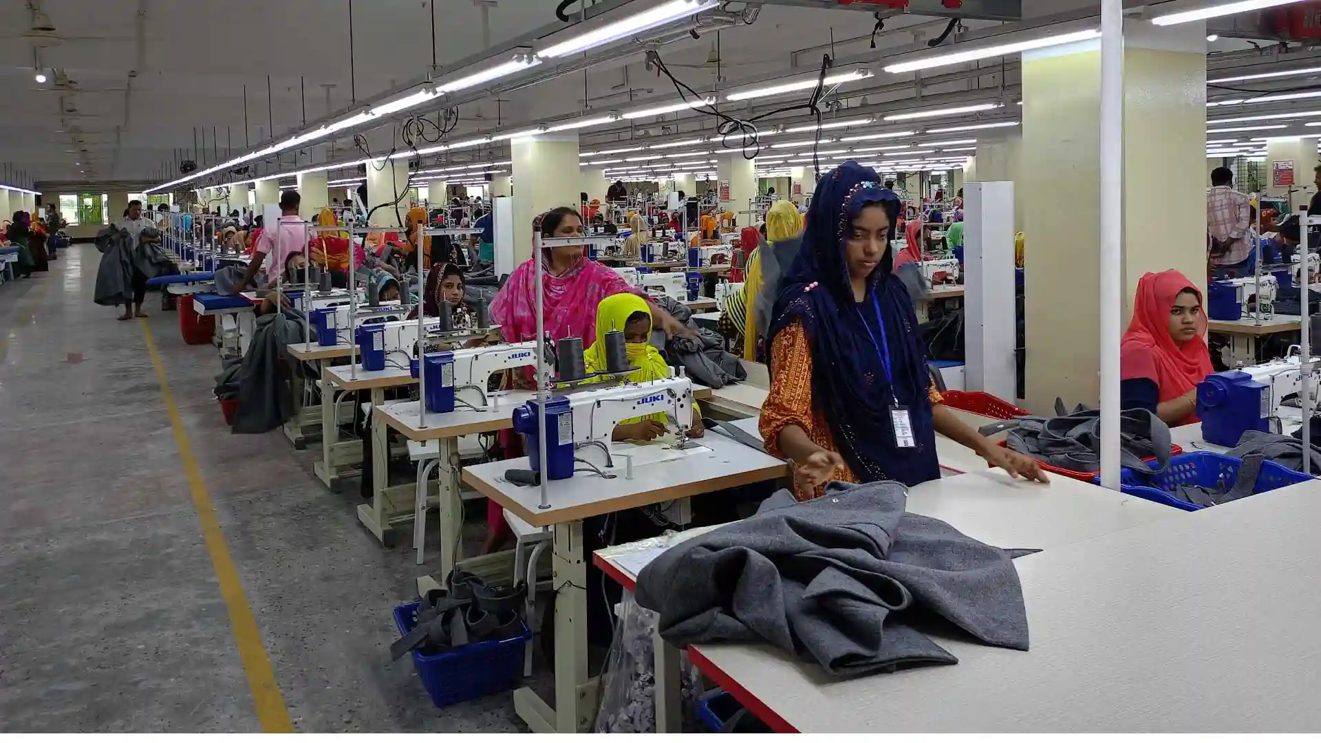 Outerwear factory production floor with workers sewing and handling garments under industrial lighting.
