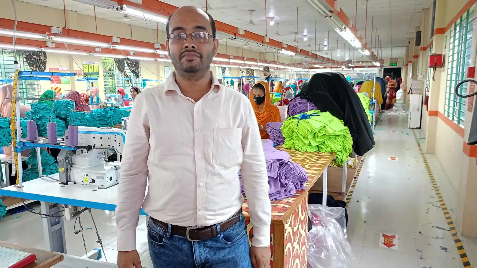 RYZEAL Managing Director visiting Raks Fashion Factory, standing inside the production floor with colorful garments stacked and workers engaged in sewing.