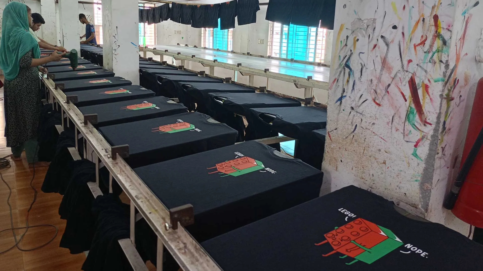Workers performing multicolor screen printing on black T-shirts in a garment printing section.